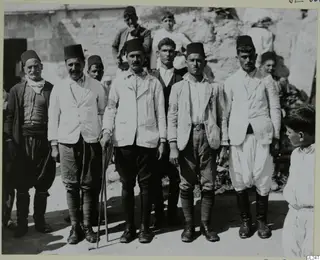 Group of men in dark brimless caps and light jackets posing outdoors at Ayia Irini, one holding a walking stick