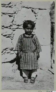 Young child in a striped smock and sash standing in front of a rough stone wall