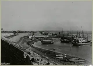Harbour at Famagusta with moored sailing boats and ships beside an old stone city wall and a railway track.
