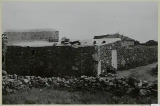 Stone building with a whitewashed flat roof and a rounded whitewashed structure on top, beside a rubble wall and dirt track.