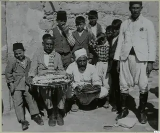 Group of men and boys posed against a plastered wall, with a seated man holding a tray of food and a kneeling bearded man beside him