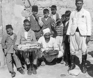 Group of men and boys posed against a plastered wall, two of the men holding trays of food
