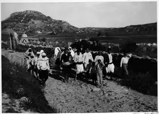 Group of people walking along a dirt track past upright stones and a small domed structure, with a rocky hill behind, at Lindros