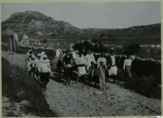 Group of people, women and men with children, walking along a sandy track past a stone wall, with a rocky hill behind.