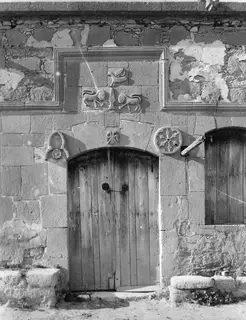 Stone building front with a wooden plank door under an arch, carved reliefs above it and a rosette medallion beside the door.