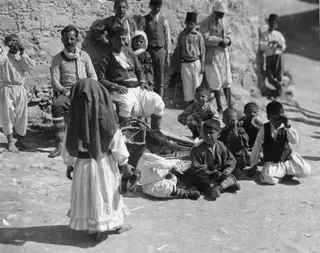 Children, men and a woman gathered by a rough stone wall, with a veiled woman standing in the foreground.