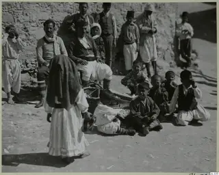 Group of children and men in front of a rough stone wall, with a woman in a dark head shawl walking in the foreground.