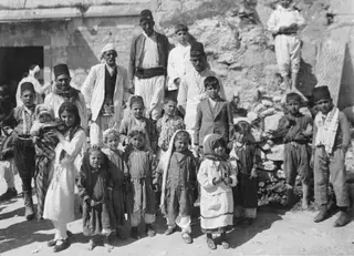 Group of men, boys and girls posing in front of a rough stone wall with a heap of stones beside it.