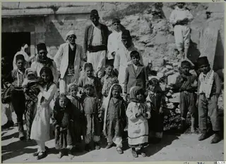 Group of people, many of them children, posed in front of a stone building and a heap of stones.