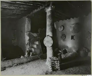 Interior of a house with a mud-plastered timber post, a raised mud bench, hanging baskets and a low stool of bundled canes.