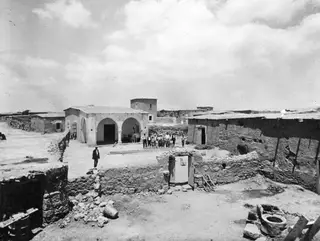 Village courtyard with arched white buildings, a stone wall and a group of people standing in the centre