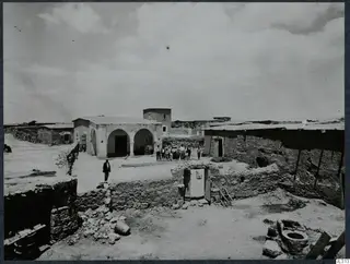 Village buildings with a whitewashed arched house, a stone wall and a group of people standing in a dusty courtyard.