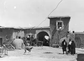 Dusty yard of a mud-brick village at Galinoporni, with a domed clay oven, a dog, and people by an arched gateway.
