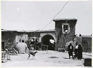 Mud-brick courtyard with a domed oven on the left, an arched gateway, a thin dog and several people