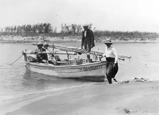 Small wooden boat in shallow water with four people, a woman stepping out onto the sandy shore, reeds behind