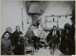 Men seated in a café interior, several smoking hookahs, with a boy on the left and a man in an apron at the stove