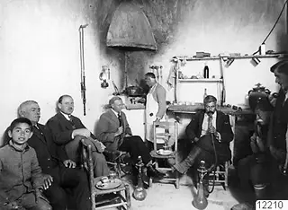 Men seated and standing in a whitewashed café interior, a boy in front, with water pipes on the floor.