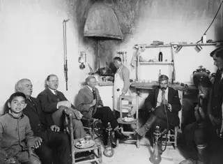 Men and a boy sit in a whitewashed café room, and a seated man at right smokes a hookah through a long hose.
