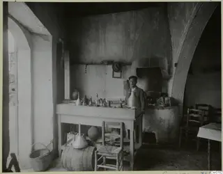 Man standing behind a wooden table covered with metal funnels, jugs and bottles in a whitewashed room with an arched doorway