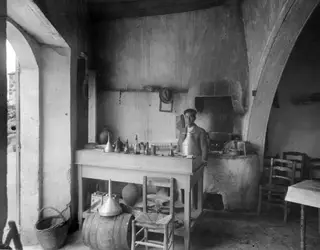 Interior of a whitewashed room with a standing man, a wooden table set with metal jugs and funnels, a barrel and a basket.