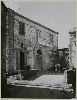 Two-storey stone building with a balustraded balcony and arched doorway, with several people on an unpaved street in Nicosia.