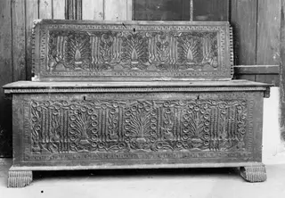 Carved wooden clothes chest with a raised panel leaning against the back, against a wooden wall.
