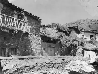 Stone houses with a wooden balcony and a low stone wall at Lazanias, with a hillside behind