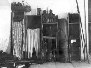 Shelved storeroom with carved wooden figures, a model boat, draped cloths and a tall carved post against a whitewashed wall