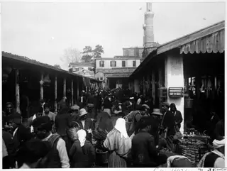 Crowded market street in Nicosia with covered wooden stalls, people with baskets, and a minaret rising behind the rooftops