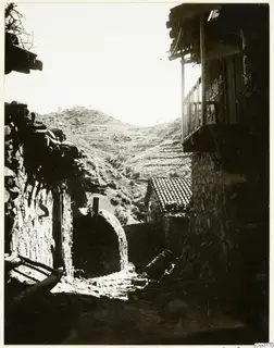 Village lane at Lazania between stone houses, with a wooden balcony on the right and a terraced hillside behind