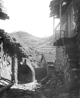 Stone-walled village lane at Lazania with a wooden balcony, tiled roofs and a hillside of terraces behind.