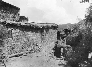 Stone-walled lane between village houses with tiled roofs at Lazanias, with mountains behind.