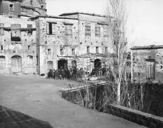 Men seated on chairs outside a café at Lapithos, under a wooden trellis in front of an old stone building