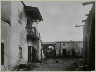 Courtyard of whitewashed houses with a balcony, an external stair, a leaning ladder and a dome-shaped oven against a wall.