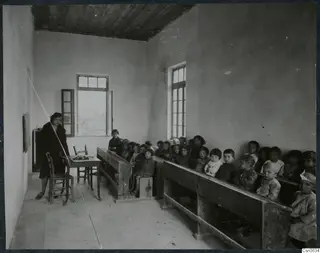 Classroom at Lapithos with a teacher standing by a table and rows of children seated on wooden benches.