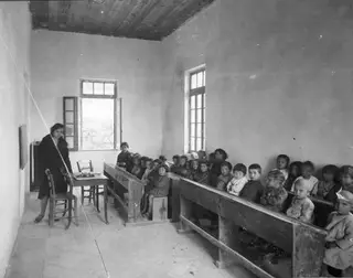 Classroom at Lapithos with a teacher standing by a small table and many children seated in rows on wooden benches.