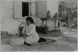Woman grinding grain with a hand mill on a pebbled yard in front of a whitewashed house wall