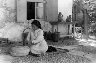 Young woman grinding grain on a hand mill in a cobbled courtyard in front of a whitewashed house wall