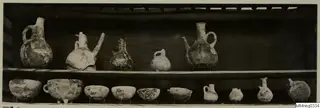 Jugs and bowls from Lapithos Markides T.9 on two shelves against a dark background.