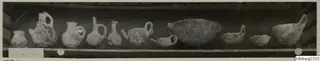 Row of about a dozen jugs and bowls from Lapithos Markides, T.4 bis A, photographed on a shelf.