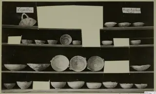 Pottery bowls and a handled jug displayed on wooden shelves, from Lapithos Markides, Tomb 1, against a dark background