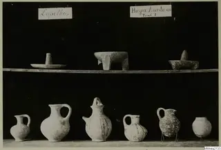 Pottery jugs and jars and small stone vessels from Lapithos, shown on two shelves against a dark background.