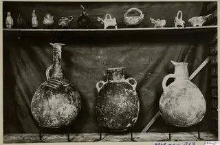 Three large pottery jars on metal stands, with small vessels on a shelf above, against a dark cloth backdrop