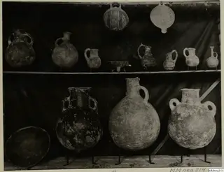 Shelved pottery vessels: small jugs and a patterned globular jar on the upper shelf, large two-handled jars on the lower shelf.