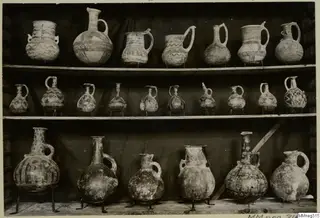Pottery jugs, juglets and jars with painted geometric patterns, arranged on three wooden shelves against a dark cloth.