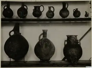 Row of small handled jugs on an upper shelf and three larger vessels on metal stands below, against a pale background