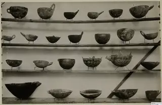 Four wooden shelves of pottery bowls and vessels with handles and spouts, some with painted patterns, each on a small wire stand.