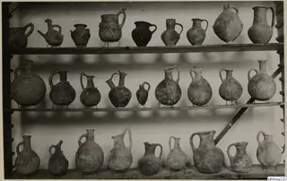 Three wooden shelves of pottery jugs and juglets, most with loop handles, a few with painted bands or patterns.