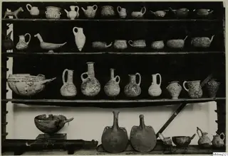 Museum shelves holding many painted pottery vessels: jugs, bowls with spouts, and two large flat-sided flasks.
