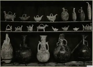 Pottery vessels on three shelves: small animal-shaped and bottle-shaped jugs above, large jars on metal stands below.